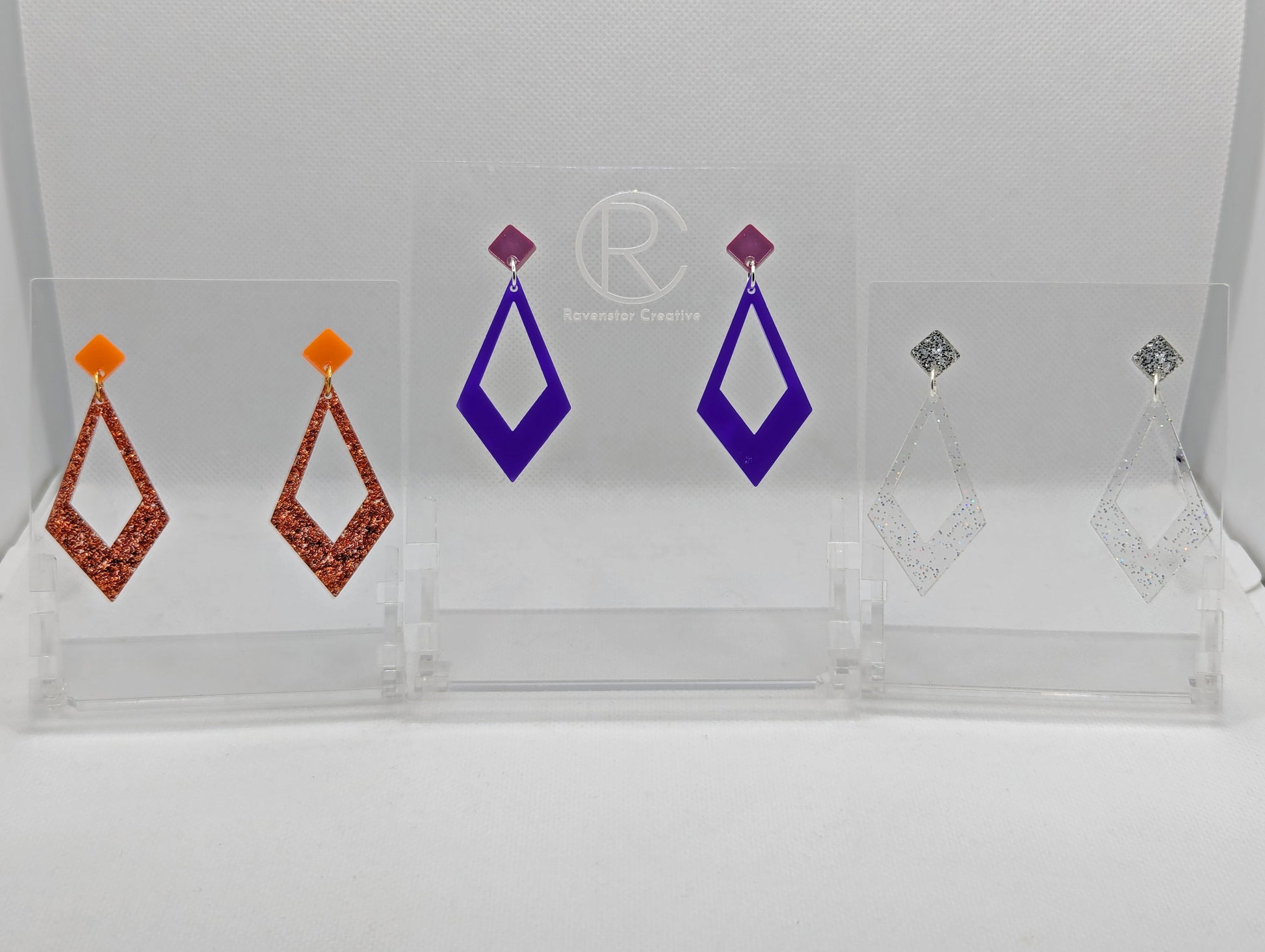 Three pairs of 2 part earrings with stud fixings. The stud is on a small diamond shape with a larger kite shape dangling under. The colours are orange top and Copper glitter bottom, grape top and purple bottom, silver glitter top and clear sparkle