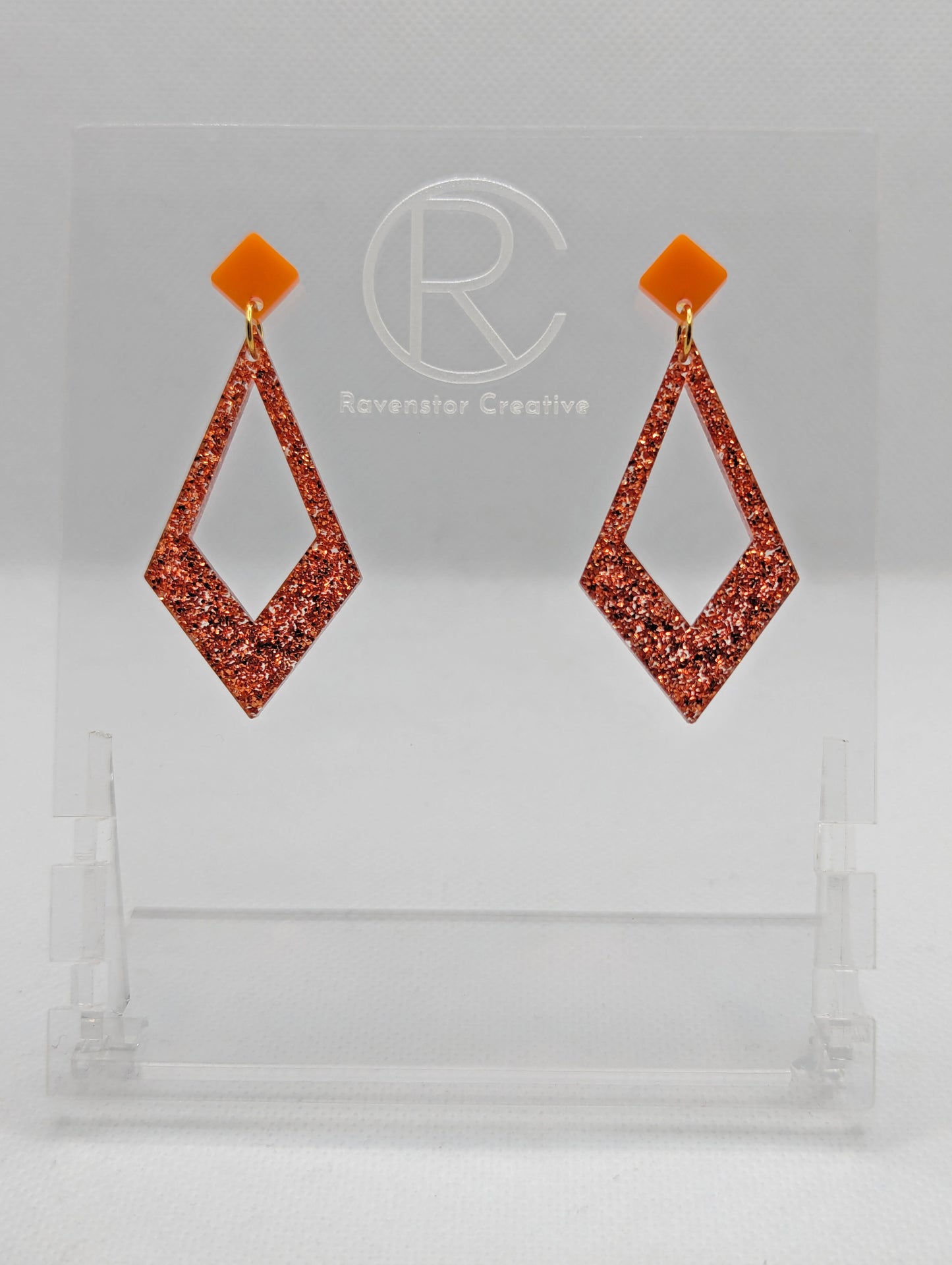 Shows a pair of 2 part earrings with stud fixings. The stud is on a small diamond shape with a larger kite shape dangling underneath. The colours are orange top and Copper Glitter Kite. They are on a clear stand against a white backing