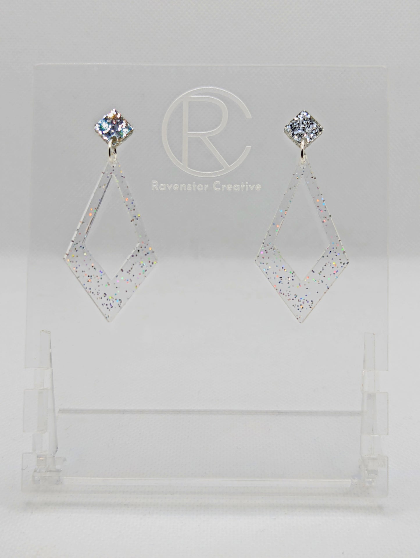 Shows a pair of 2 part earrings with stud fixings. The stud is on a small diamond shape with a larger kite shape dangling underneath. The colours are silver glitter top and clear sparkle Kite. They are on a clear stand against a white backing