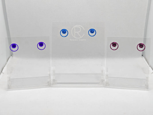 Shows three pairs of small studded acrylic earrings that are a circle with another circle around it joining at the top. The colours are Purple, Royal Blue Glitter and Pink Glitter. They are against a white background.