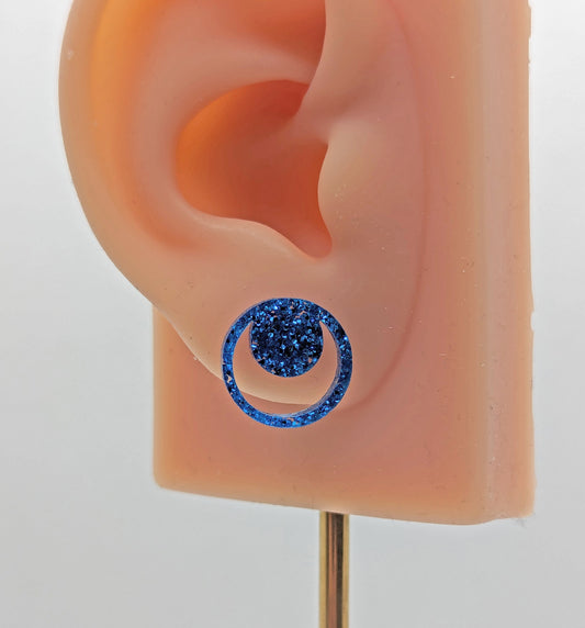 Shows a pair of small studded acrylic earrings that are a circle with another circle around it joining at the top. They are Royal Blue Glitter in colour. They are on an ear to show the size.