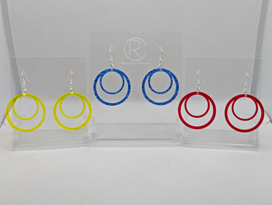 Shows three pairs of earrings that have ear wire hook fixings. They are large double hoops joined at the top. The colours are Neon Yellow, Royal Blue Glitter and Translucent Red. They are against a white background on see through stands.