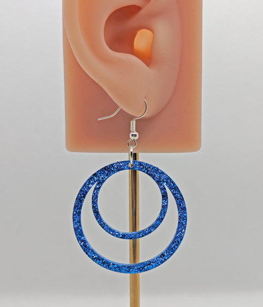 Image of a pair of large double hoop earrings joined at the top on an ear showing the size against a white background. They are royal blue glitter in colour and have silver fixings.