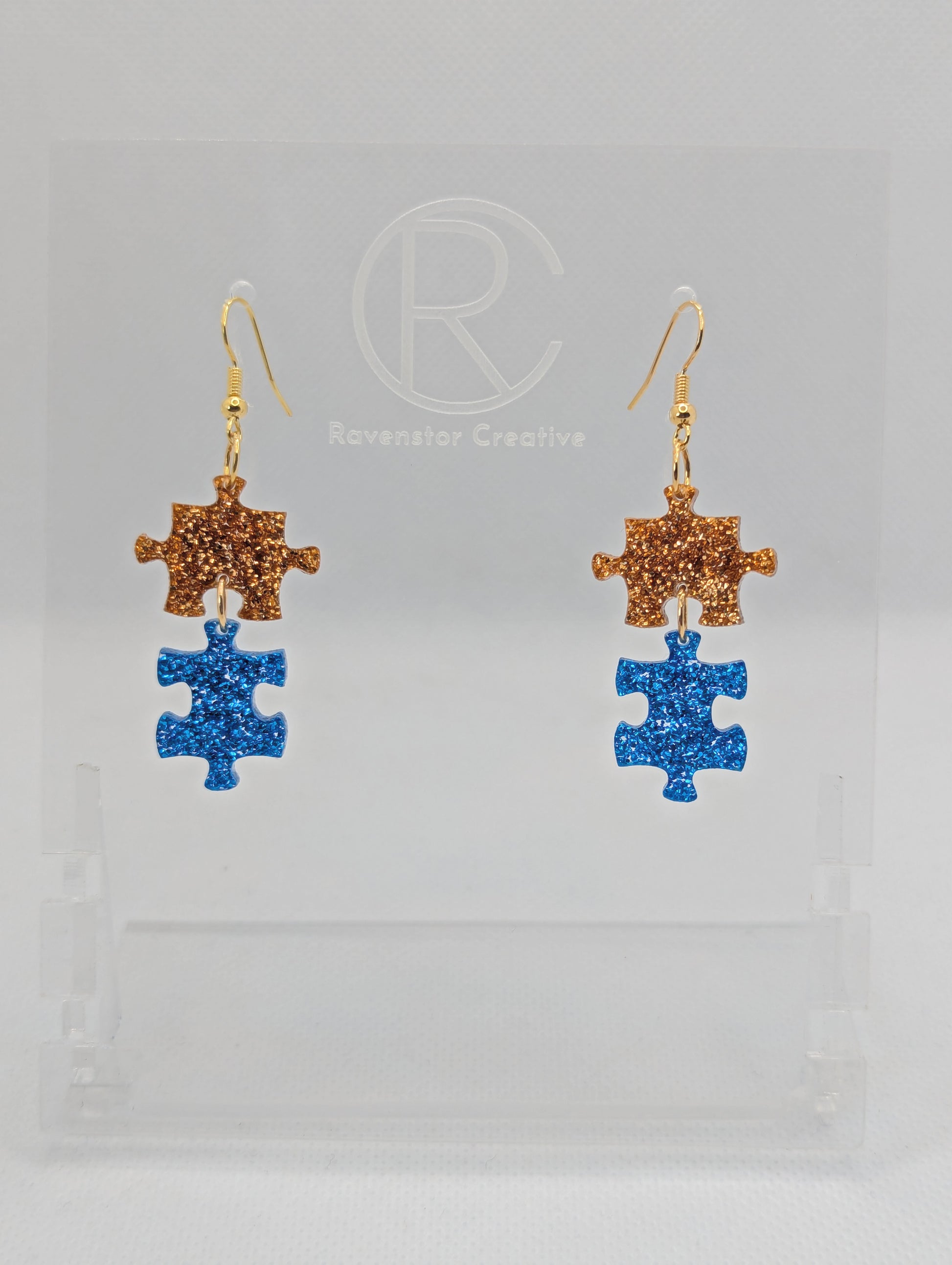 Shows a pair of acrylic earrings with gold ear wire hook fixings. They are Two Jigsaw Game Pieces with one dangling underneath. The colours are copper glitter and royal blue glitter. They are against a white background on a clear stand.