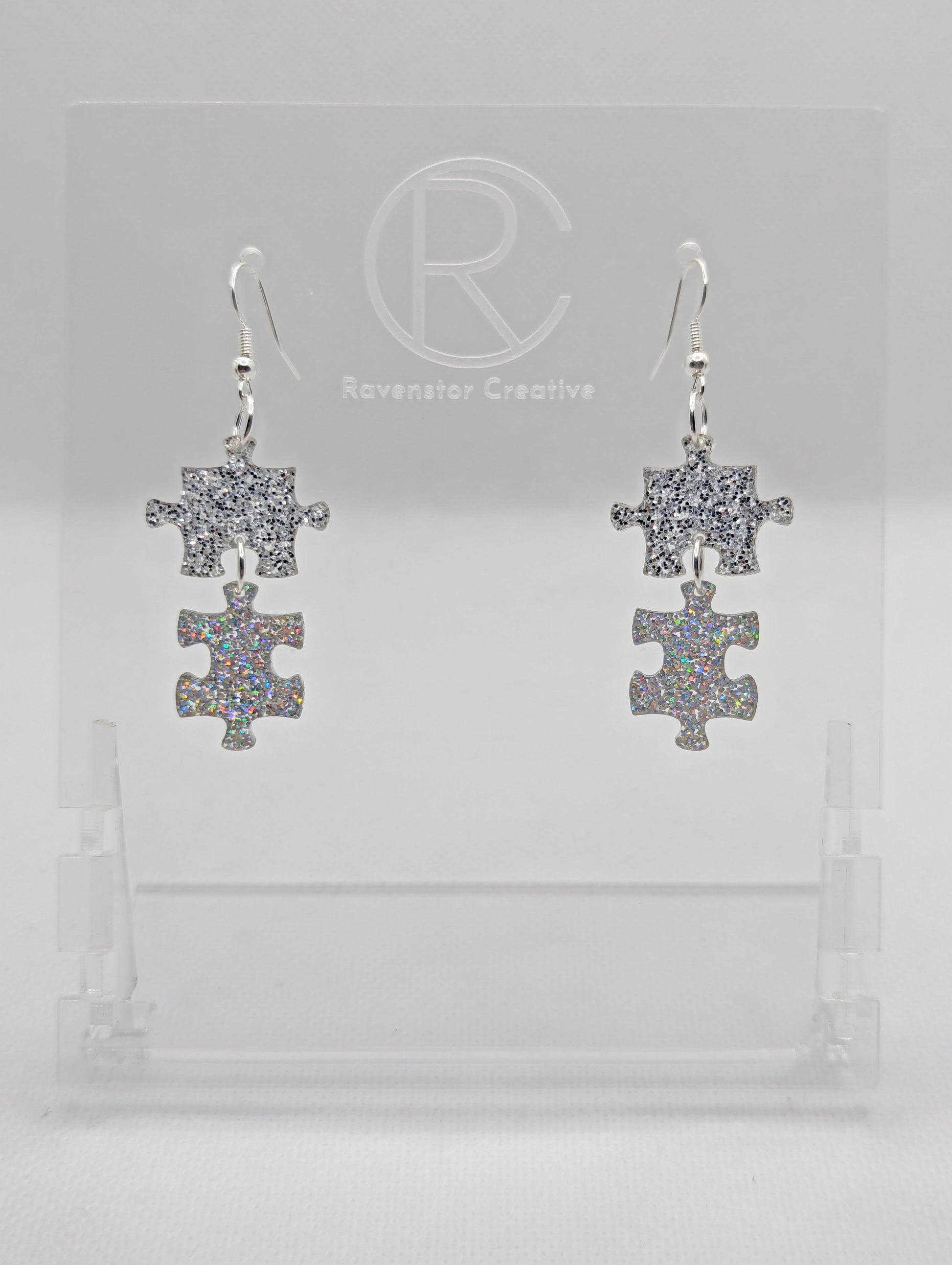 Shows a pair of acrylic earrings with silver ear wire hook fixings. They are Two Jigsaw Game Pieces with one dangling underneath. The colours are Silver glitter and Holographic Glitter. They are against a white background on a clear stand.