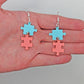 Shows a pair of acrylic earrings with silver ear wire hook fixings. They are Two Jigsaw Game Pieces with one dangling underneath. They are against a hand showing the size of Two finger widths in length and one wide.