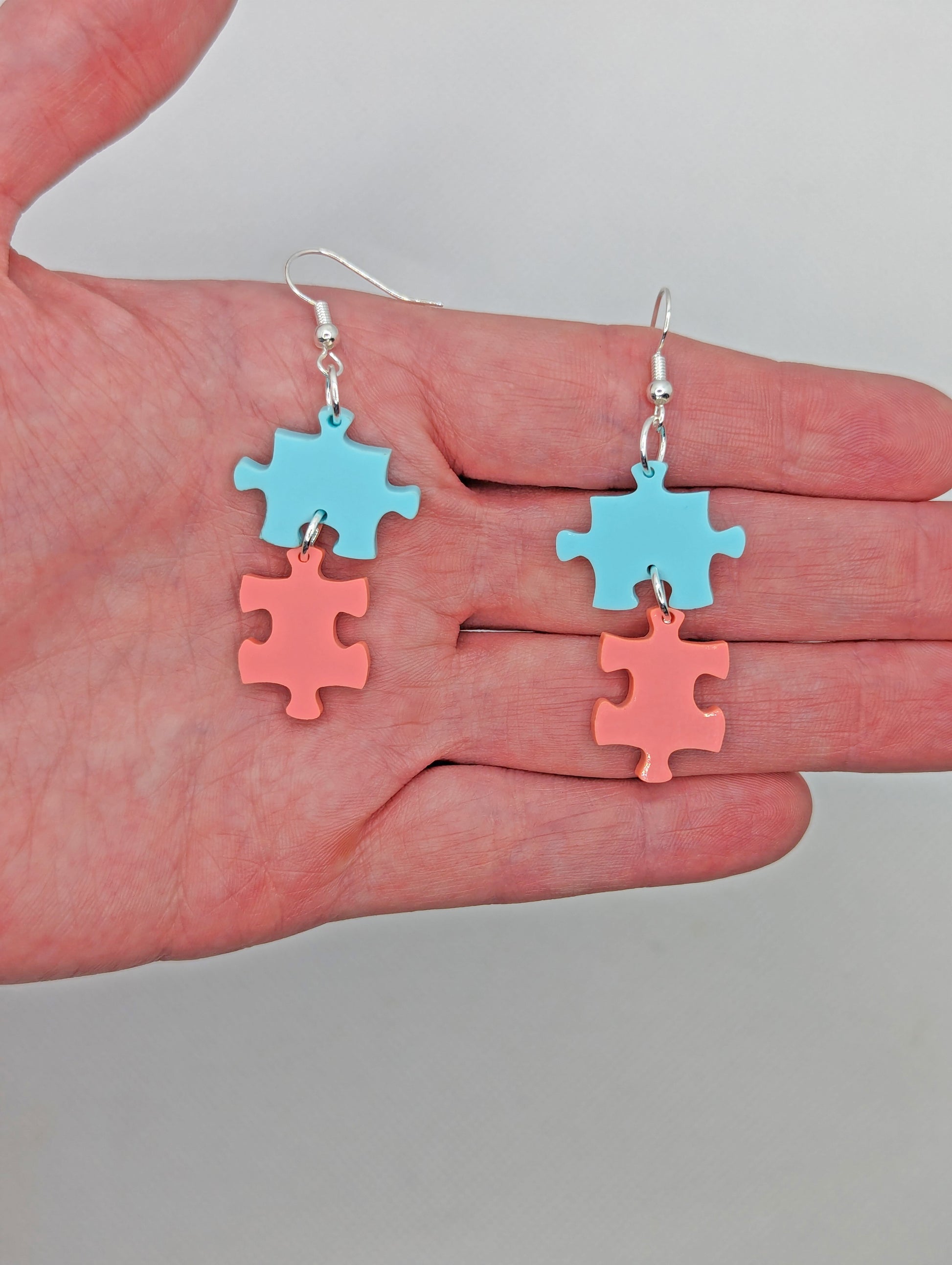 Shows a pair of acrylic earrings with silver ear wire hook fixings. They are Two Jigsaw Game Pieces with one dangling underneath. They are against a hand showing the size of Two finger widths in length and one wide.