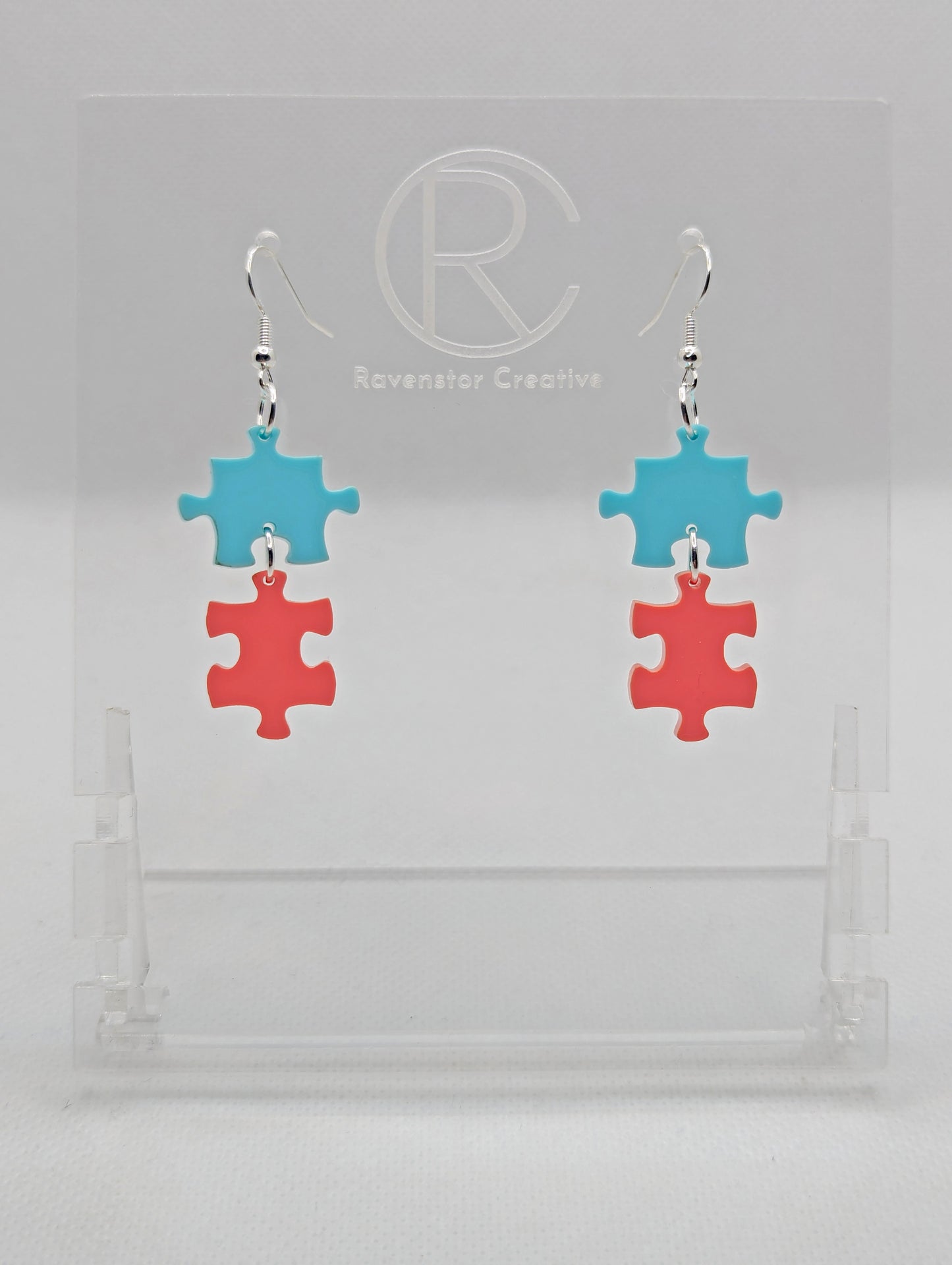 Shows a pair of acrylic earrings with silver ear wire hook fixings. They are Two Jigsaw Game Pieces with one dangling underneath. The colours are Spearmint and Sherbet Pink. They are against a white background on a clear stand.