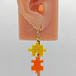 Shows an acrylic earring with silver ear wire hook fixings. It is Two Jigsaw Game Pieces with one dangling underneath. They are Yellow and Orange in colour. It is on an ear to show the size.