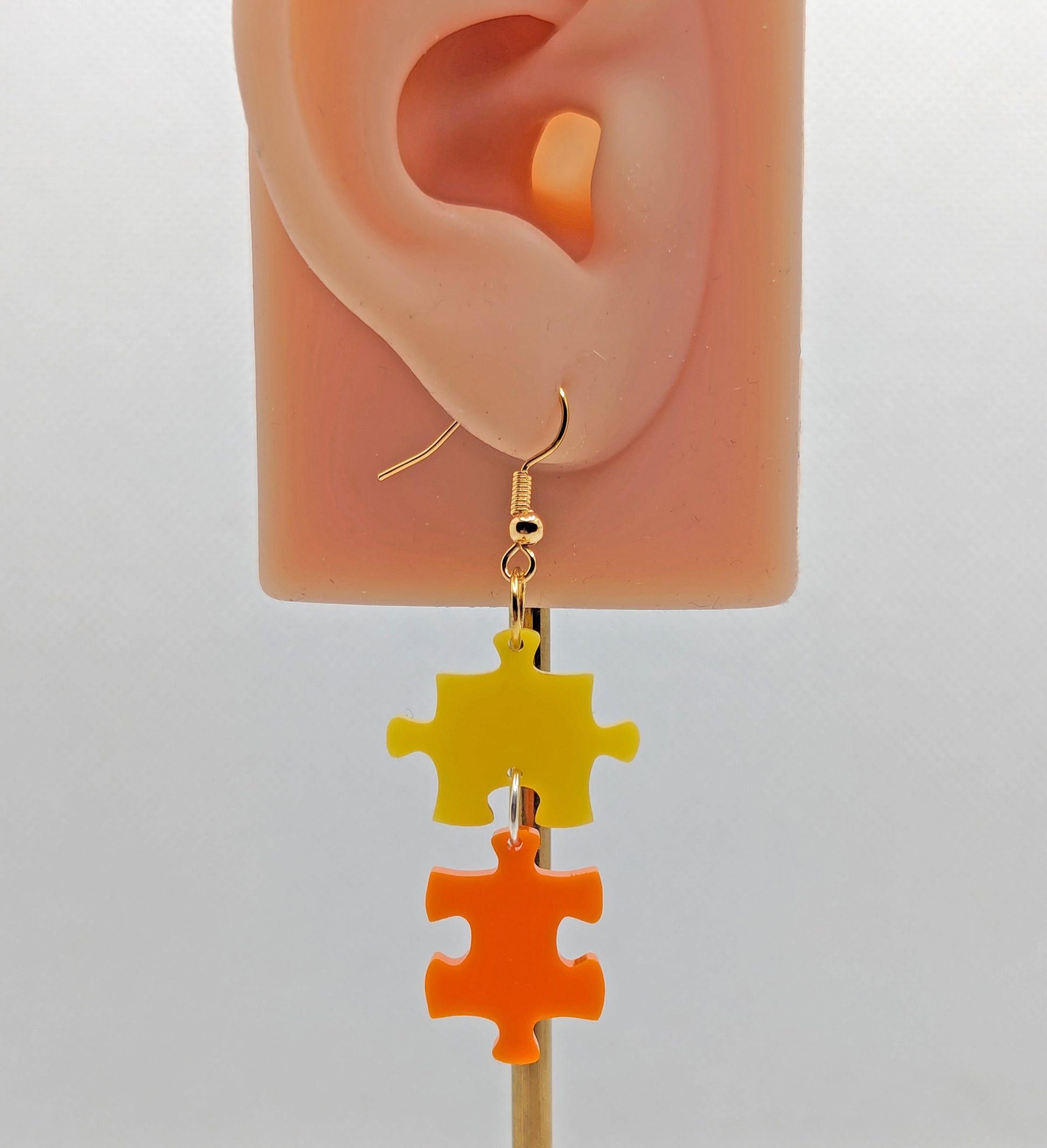 Shows an acrylic earring with silver ear wire hook fixings. It is Two Jigsaw Game Pieces with one dangling underneath. They are Yellow and Orange in colour. It is on an ear to show the size.