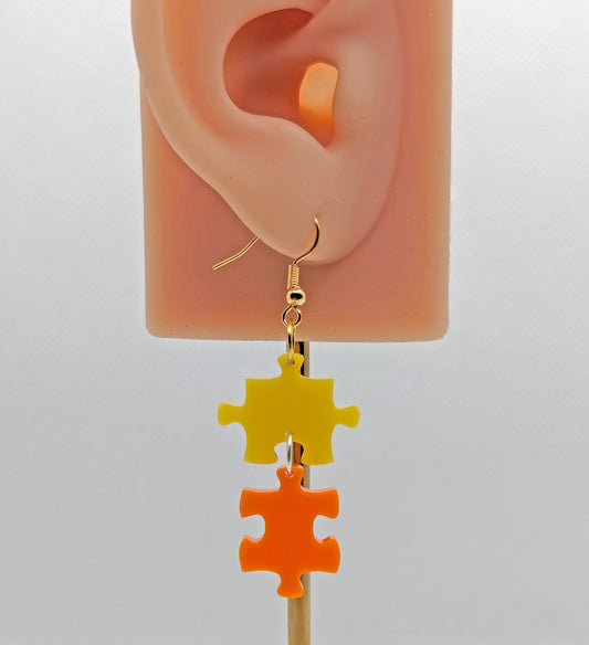 Shows an acrylic earring with silver ear wire hook fixings. It is Two Jigsaw Game Pieces with one dangling underneath. They are Yellow and Orange in colour. It is on an ear to show the size.