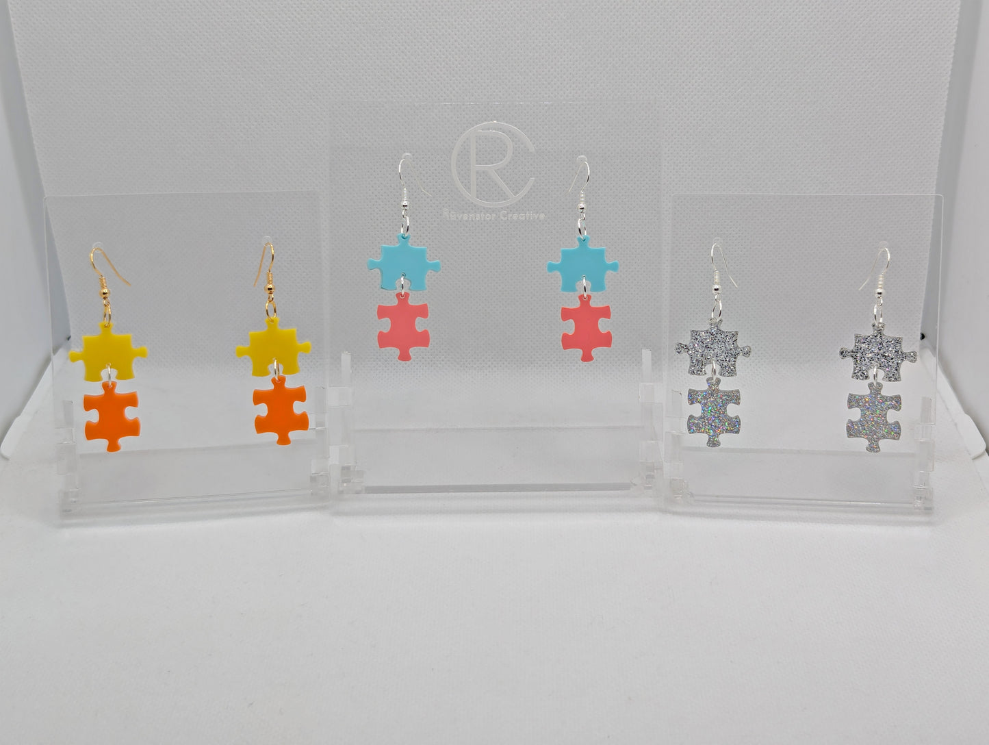 Shows three pairs of acrylic earrings with ear wire hook fixings. They are Two Jigsaw Game Pieces with one dangling underneath. The colours are Yellow and Orange, Spearmint and Sherbet Pink, Silver and Holographic Glitter.