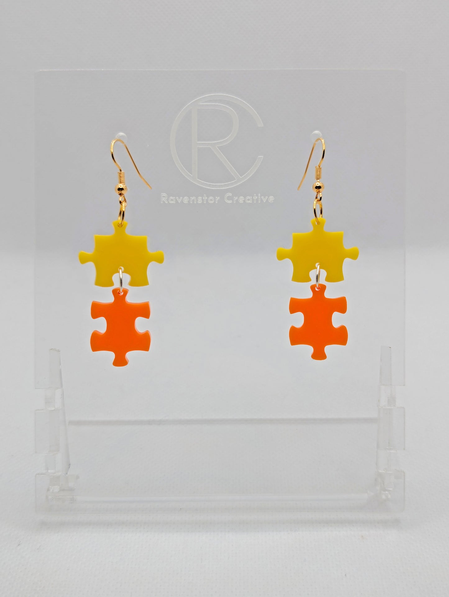 Shows a pair of acrylic earrings with silver ear wire hook fixings. They are Two Jigsaw Game Pieces with one dangling underneath. The colours are Yellow and Orange. They are against a white background on a clear stand.