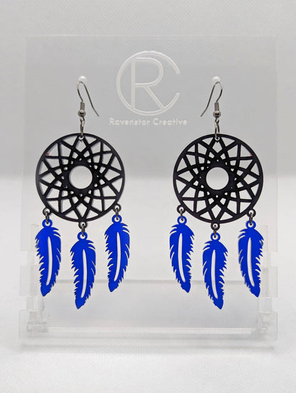 A pair of large earrings shaped like a dreamcatcher. A patterned circle at the top with 3 dangling shapes under. They are made from acrylic and are black for the circle and translucent Blue for the dangles. They have black ear wire hook fixings.