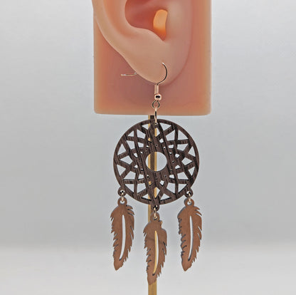 Shows earrings shaped like dreamcatchers made in two parts against an ear to show the size. A patterned circle at the top with 3 dangling feathers under. It is made from walnut and cherry woods.