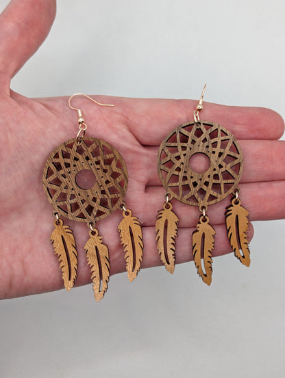 Shows earrings shaped like dreamcatchers made in two parts against a hand to show the size. A patterned circle at the top with 3 dangling shapes under. it is 4 finger widths in length and two and a half wide.  It is made from walnut and cherry wood.