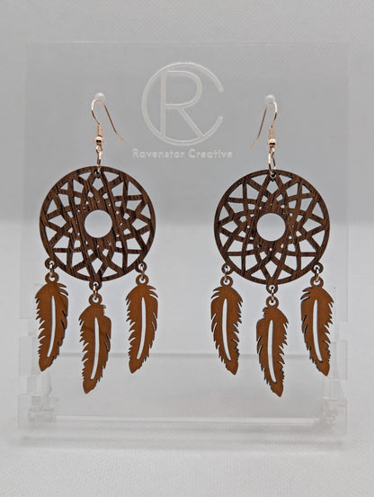 A pair of large earrings shaped like a dreamcatcher. A patterned circle at the top with 3 dangling shapes under. They are made from walnut and Cherry wood and have gold ear wire hook fixings. They are on a clear stand against a white background.