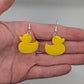 Image of a pair of rubber ducky / duck shaped earrings on a hand showing the size against a white background, they are one and a half finger widths length  and the same again wide. They are yellow and have silver fixings.