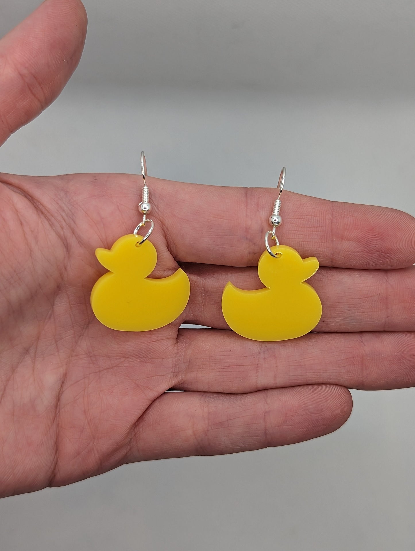 Image of a pair of rubber ducky / duck shaped earrings on a hand showing the size against a white background, they are one and a half finger widths length  and the same again wide. They are yellow and have silver fixings.