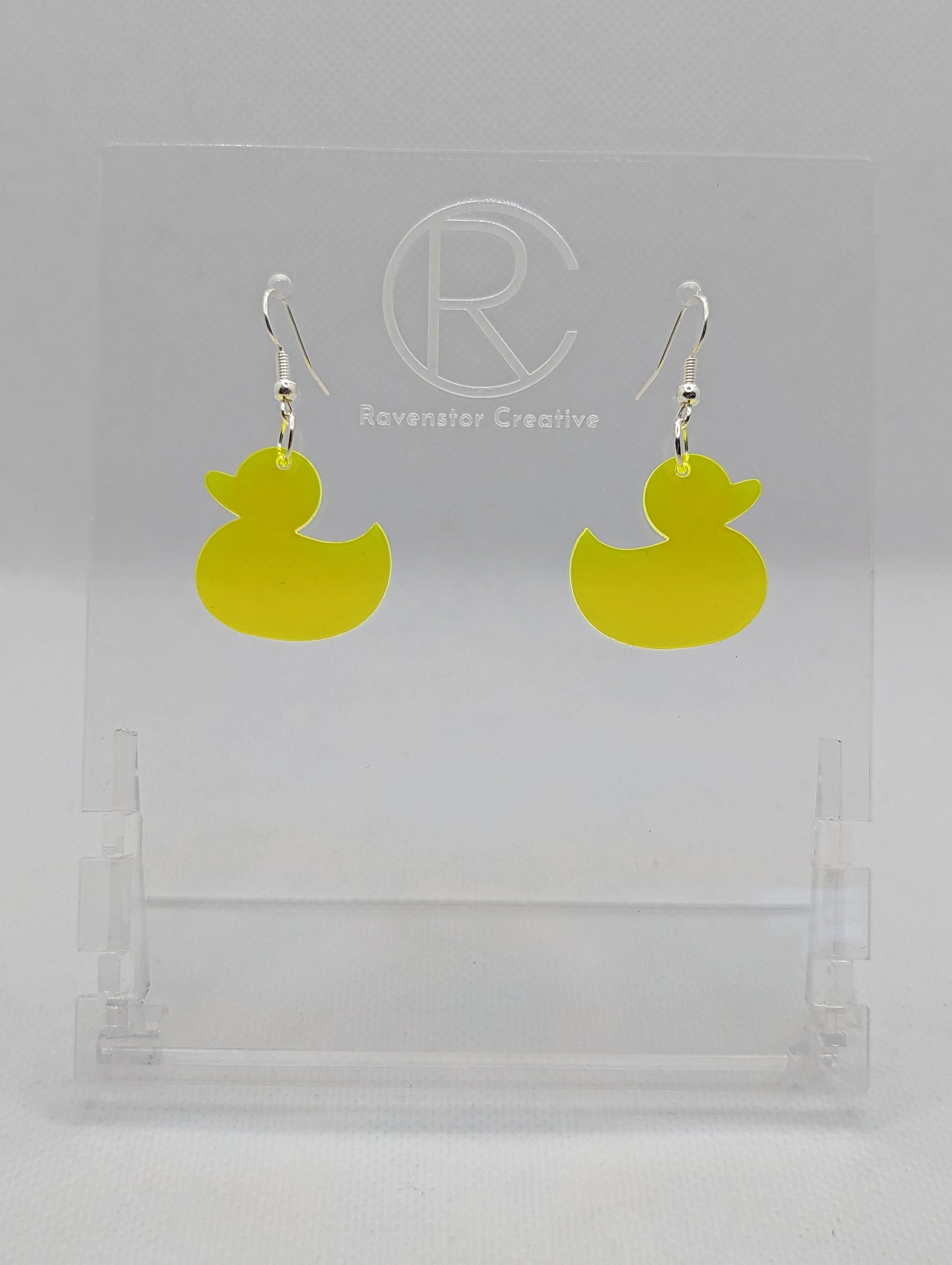 Shows a pair of acrylic earrings with silver ear wire hook fixings. They are rubber ducky / duck shapes. The colour of them is neon orange. They are on a see through stand that has the Ravenstor Creative Logo and its against a white background.