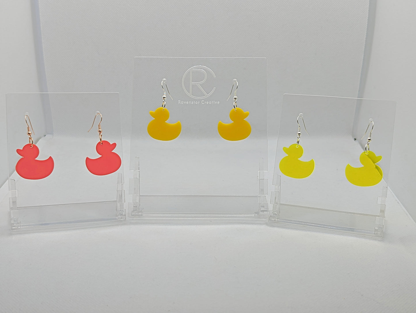 Shows three pairs of earrings that have ear wire hook fixings. They are rubber Duck / ducky shaped with ear wire hook fixings. The colours are Neon pink, yellow and neon Yellow. They are against a white background on see through stands.