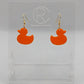 Shows a pair of acrylic earrings with gold ear wire hook fixings. They are rubber ducky / duck shapes. The colour of them is orange. They are on a see through stand that has the Ravenstor Creative Logo and its against a white background.