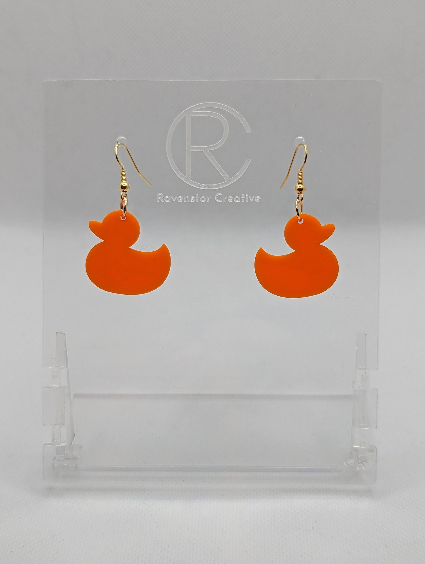 Shows a pair of acrylic earrings with gold ear wire hook fixings. They are rubber ducky / duck shapes. The colour of them is orange. They are on a see through stand that has the Ravenstor Creative Logo and its against a white background.