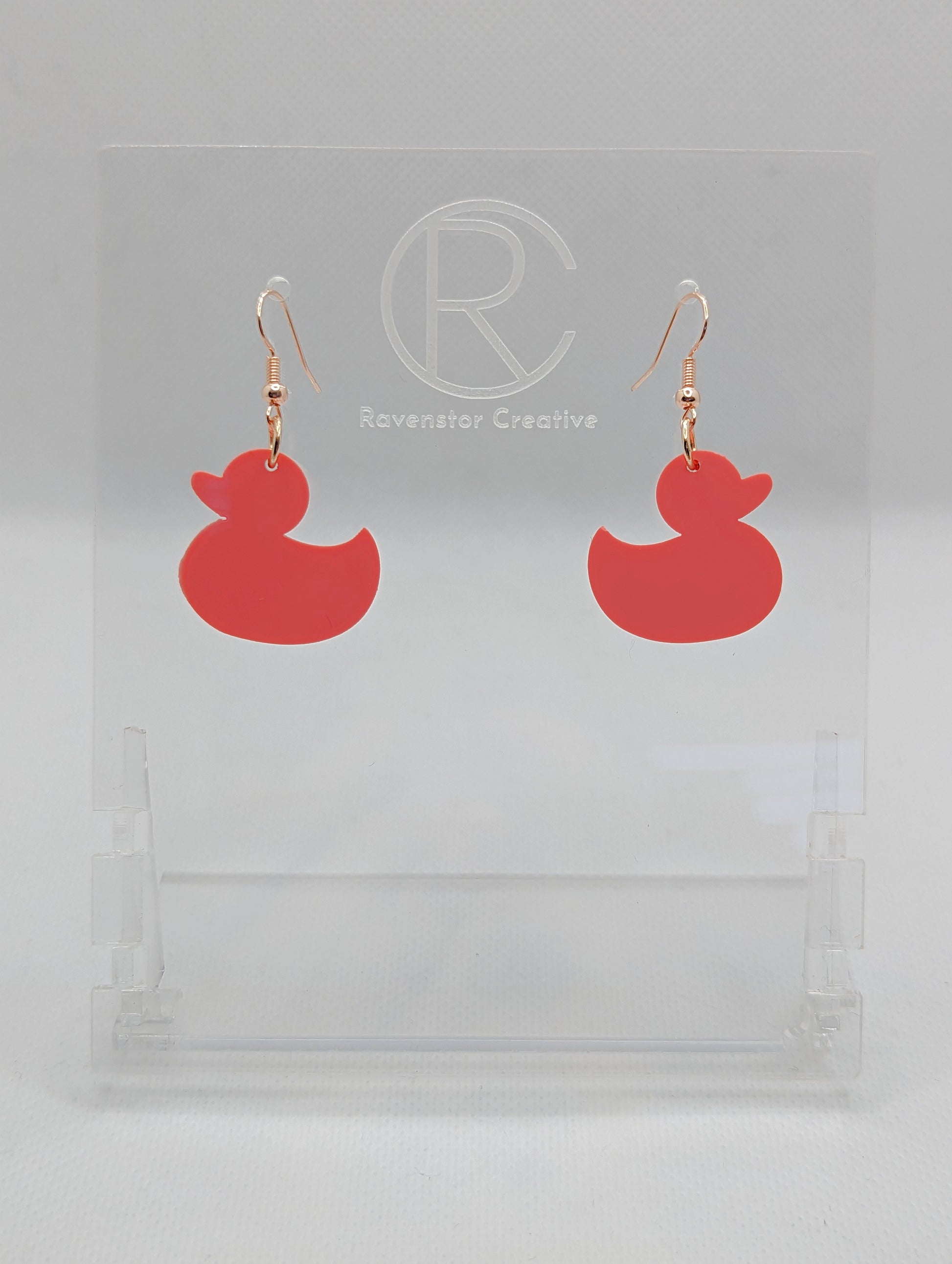 Shows a pair of acrylic earrings with gold ear wire hook fixings. They are rubber ducky / duck shapes. The colour of them is orange. They are on a see through stand that has the Ravenstor Creative Logo and its against a white background.