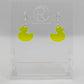 Shows a pair of acrylic earrings with silver ear wire hook fixings. They are rubber ducky / duck shapes. The colour of them is neon yellow. They are on a see through stand that has the Ravenstor Creative Logo and its against a white background.