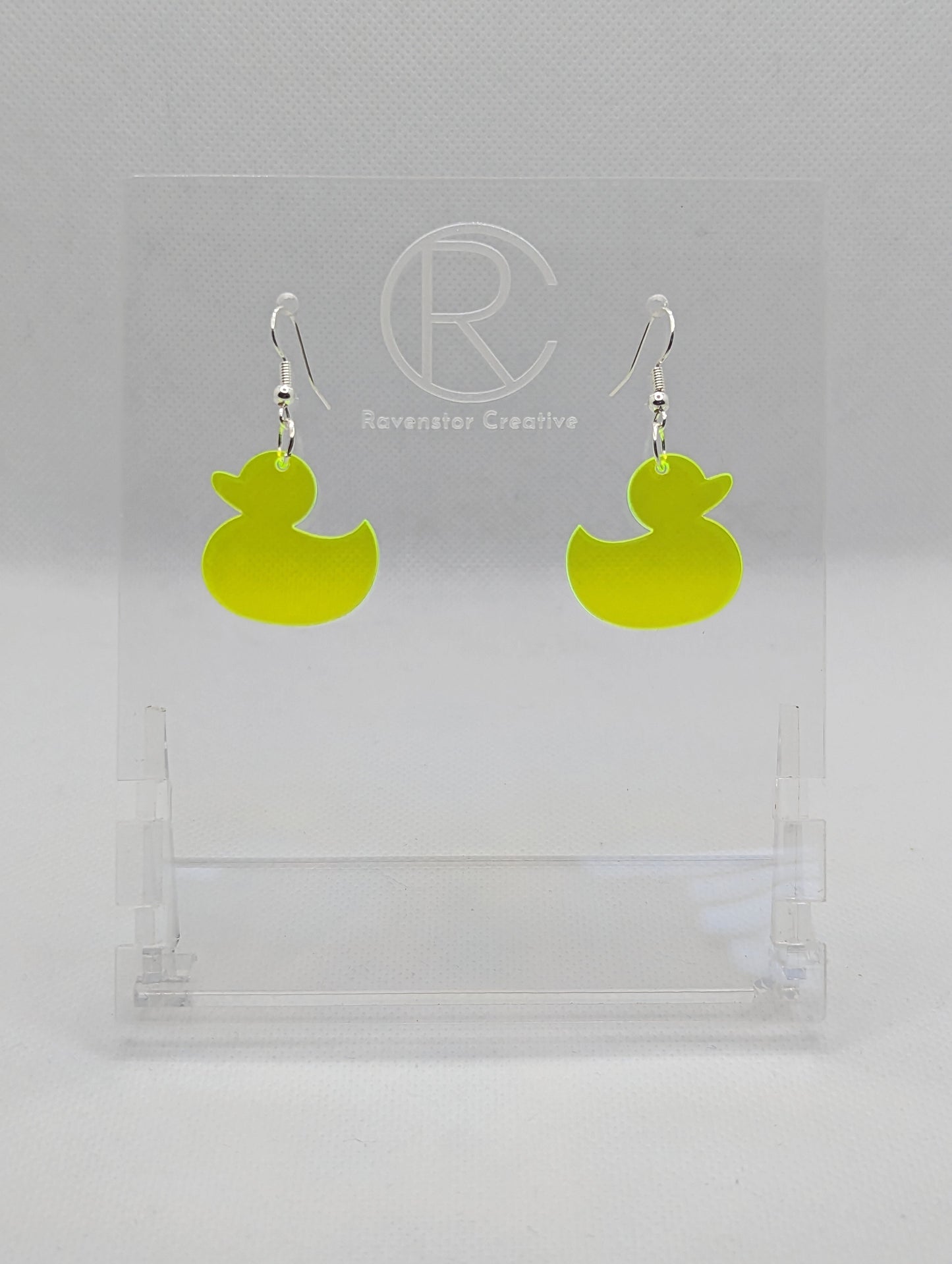 Shows a pair of acrylic earrings with silver ear wire hook fixings. They are rubber ducky / duck shapes. The colour of them is neon yellow. They are on a see through stand that has the Ravenstor Creative Logo and its against a white background.