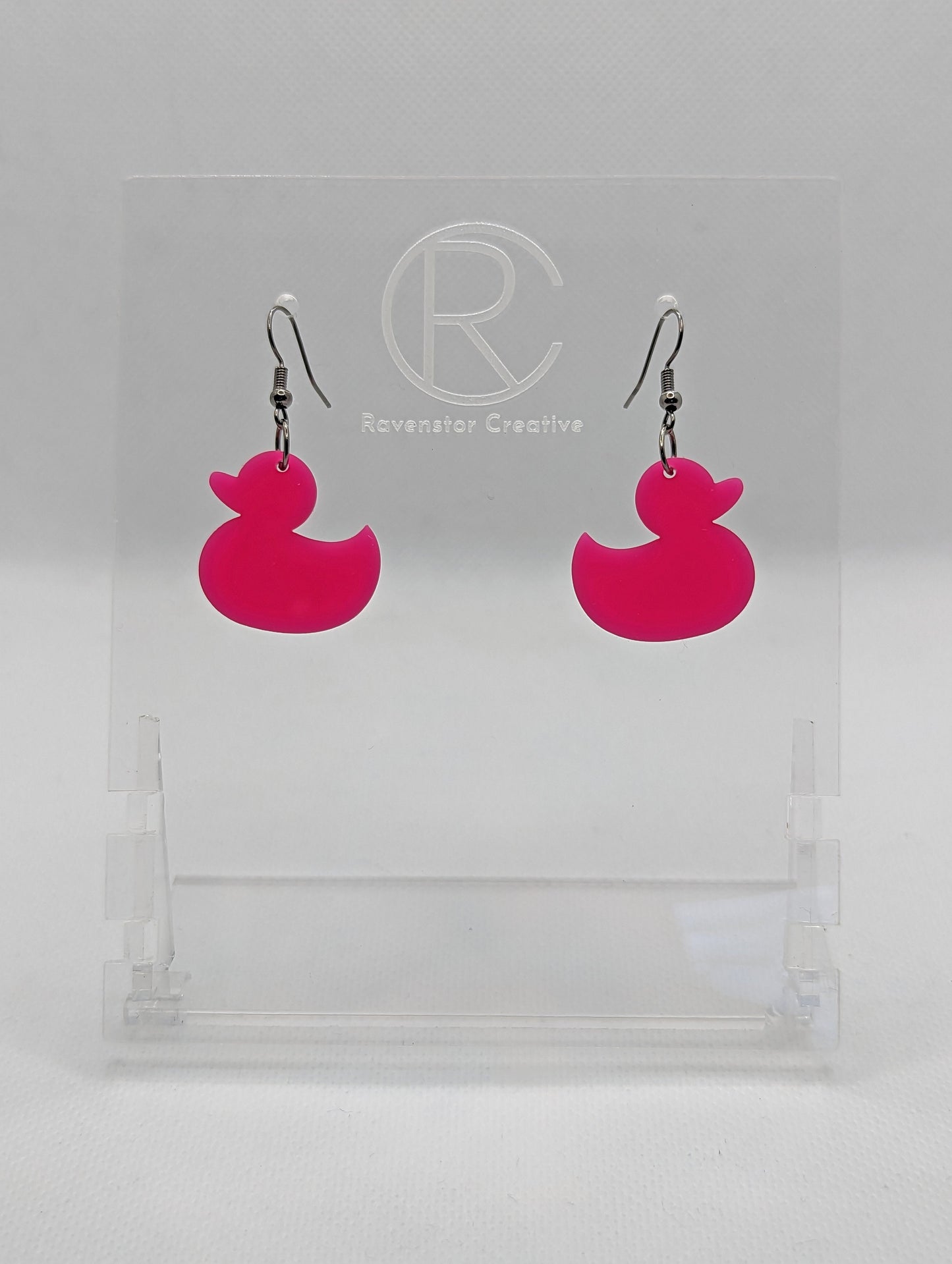 Shows a pair of acrylic earrings with black ear wire hook fixings. They are rubber ducky / duck shapes. The colour of them is hot pink. They are on a see through stand that has the Ravenstor Creative Logo and its against a white background.