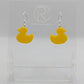 Shows a pair of acrylic earrings with silver ear wire hook fixings. They are rubber ducky / duck shapes. The colour of them is yellow. They are on a see through stand that has the Ravenstor Creative Logo and its against a white background.