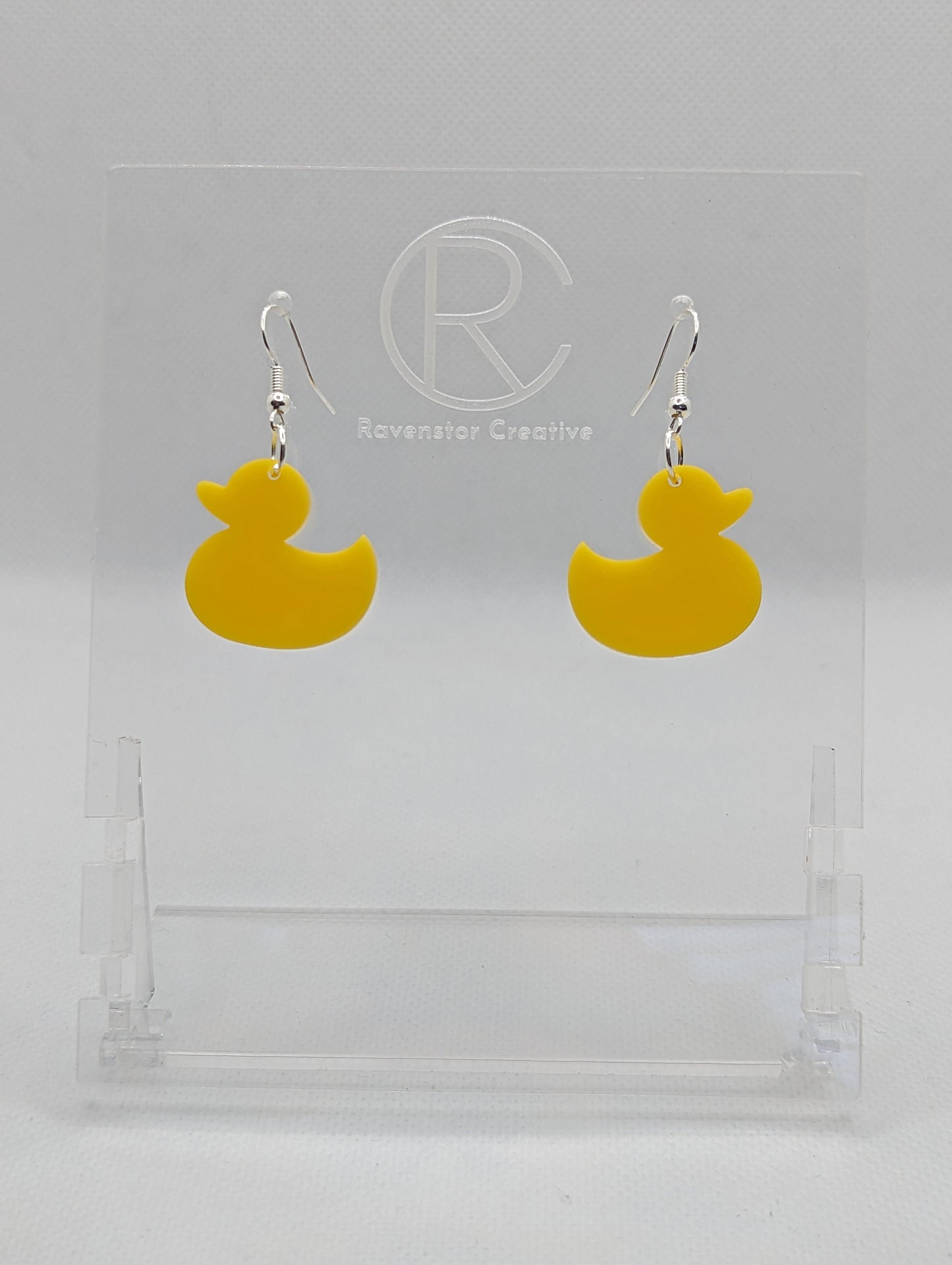 Shows a pair of acrylic earrings with silver ear wire hook fixings. They are rubber ducky / duck shapes. The colour of them is yellow. They are on a see through stand that has the Ravenstor Creative Logo and its against a white background.