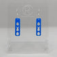 A pair of acrylic earrings that have Silver ear wire hook fixings. They are slim long bar pendant earrings with three Easter egg and rabbit / bunny shapes cut out. They are Blue in colour and are against a white background on see through stand.