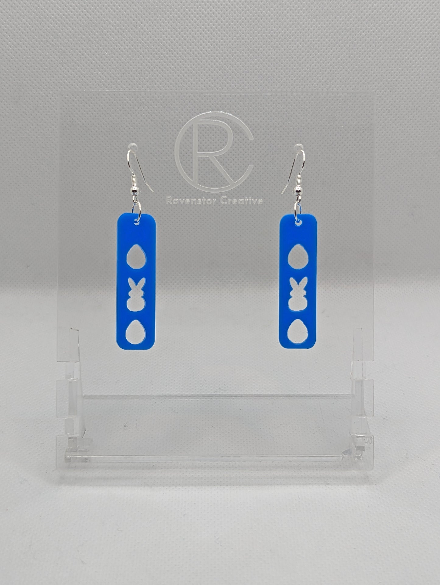 A pair of acrylic earrings that have Silver ear wire hook fixings. They are slim long bar pendant earrings with three Easter egg and rabbit / bunny shapes cut out. They are Blue in colour and are against a white background on see through stand.