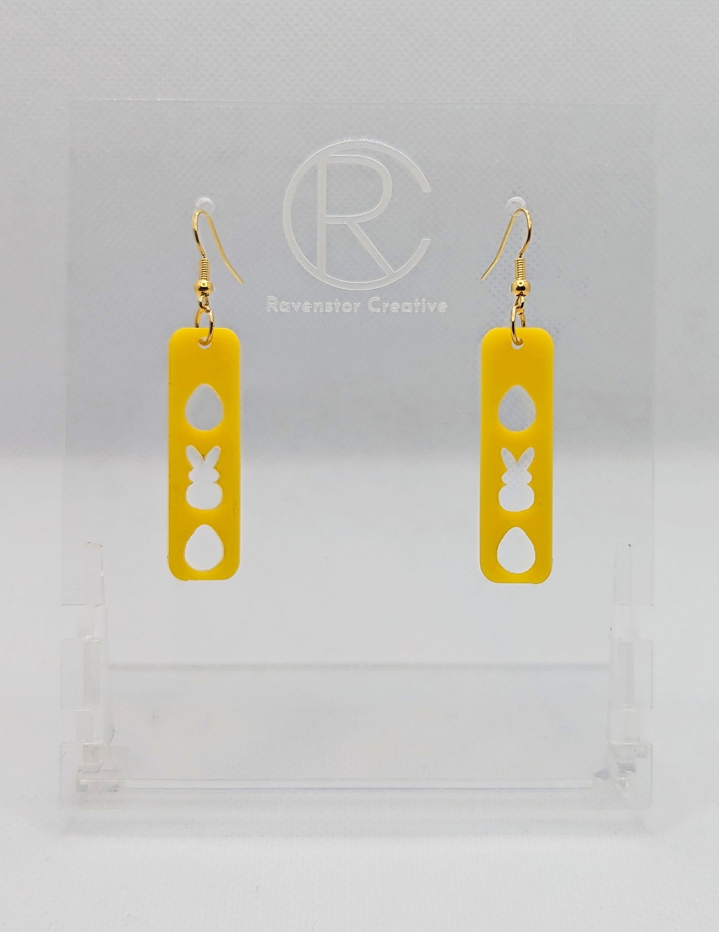 A pair of acrylic earrings that have Rose Gold ear wire hook fixings. They are slim long bar pendant earrings with three Easter egg and rabbit / bunny shapes cut out. They are Yellow in colour and are against a white background on see through stand.