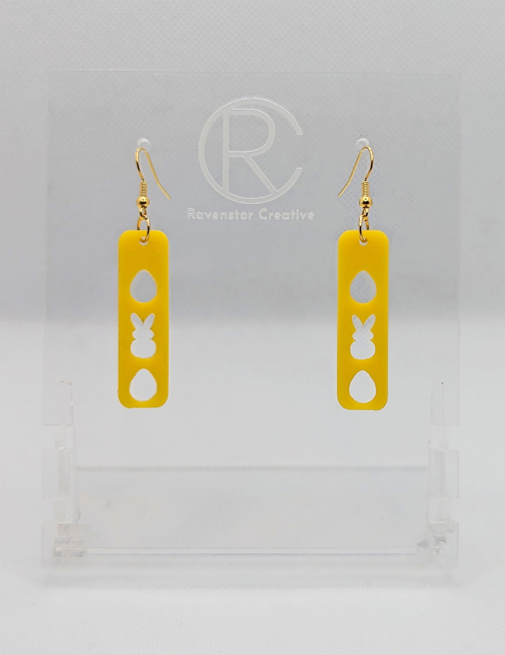 A pair of acrylic earrings that have Rose Gold ear wire hook fixings. They are slim long bar pendant earrings with three Easter egg and rabbit / bunny shapes cut out. They are Yellow in colour and are against a white background on see through stand.