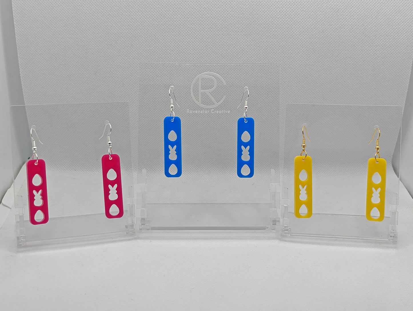 Three pairs of earrings that have ear wire fixings. They are slim long bar pendant earrings with three Easter egg and rabbit / bunny shapes cut out. The colours are hot pink, Blue and Yellow. They are against a white background on see through stands