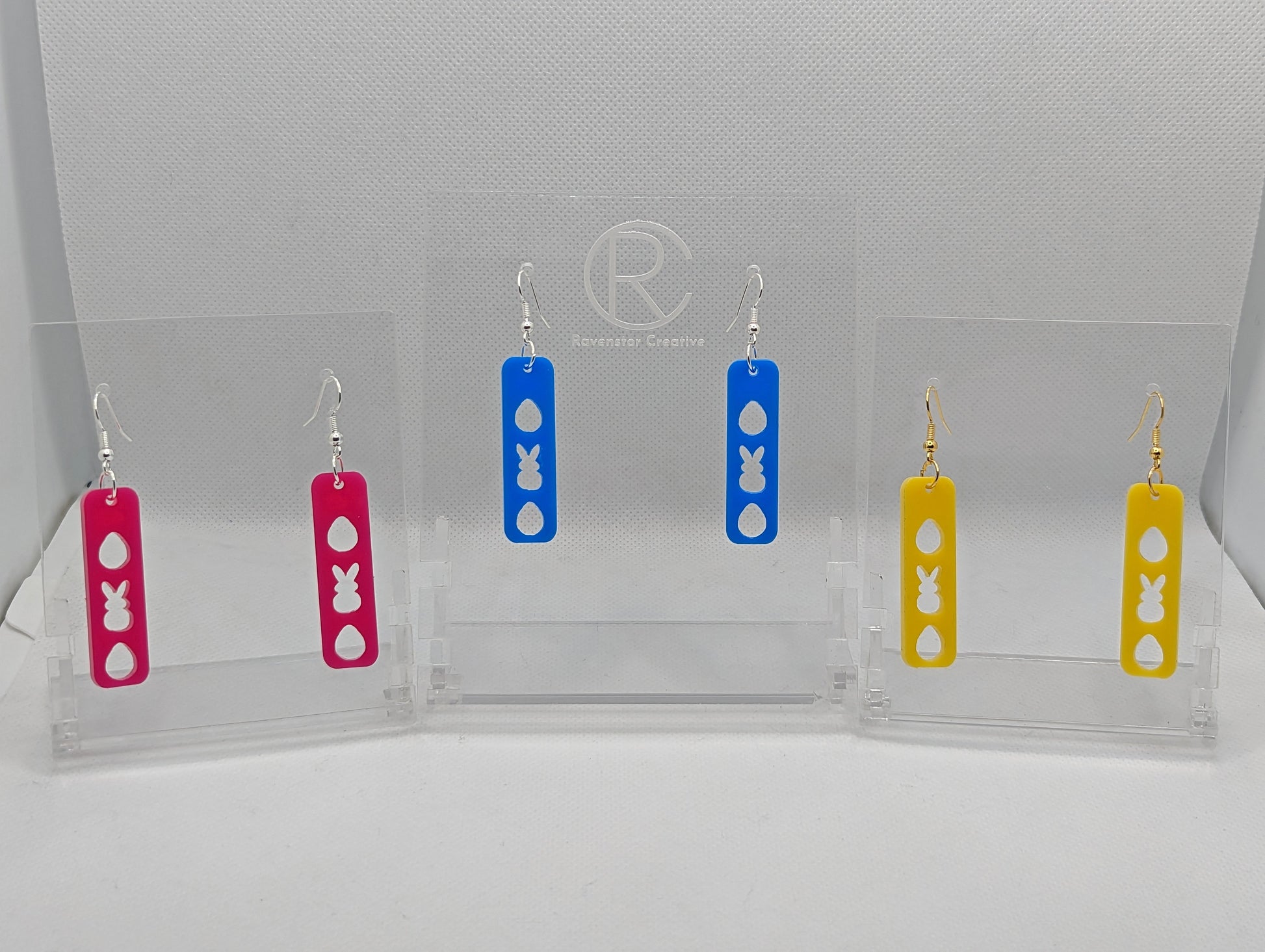 Three pairs of earrings that have ear wire fixings. They are slim long bar pendant earrings with three Easter egg and rabbit / bunny shapes cut out. The colours are hot pink, Blue and Yellow. They are against a white background on see through stands