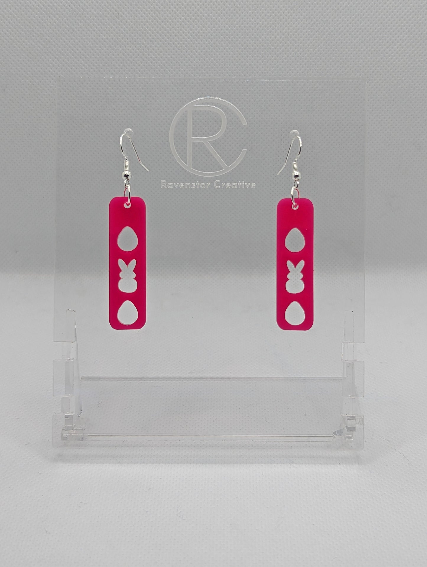 A pair of acrylic earrings that have Silver ear wire hook fixings. They are slim long bar pendant earrings with three Easter egg and rabbit / bunny shapes cut out. They are Hot Pink in colour and are against a white background on see through stand.