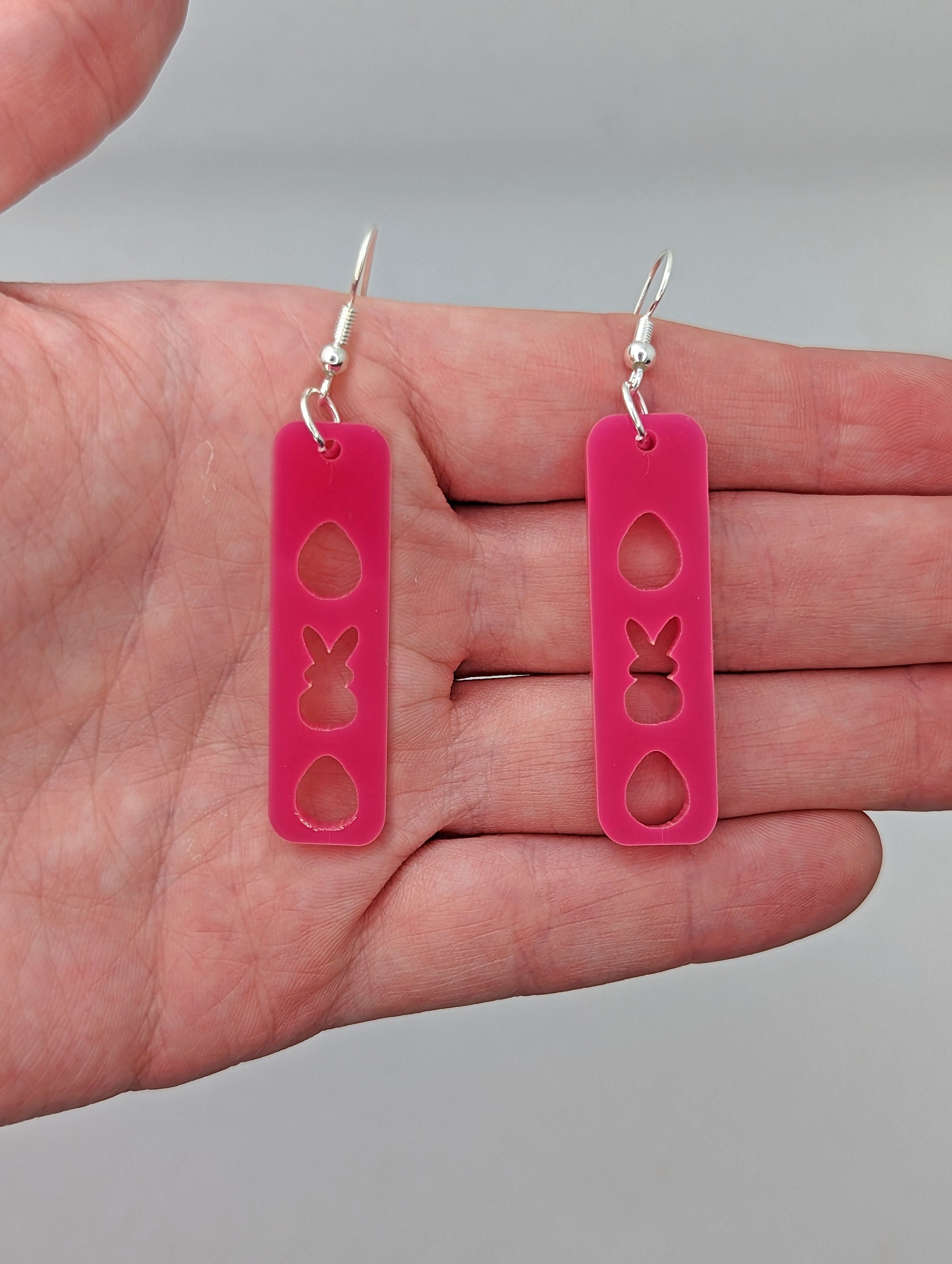 A pair of earrings that have silver ear wire fixings. They are slim long bar pendant earrings with three Easter egg and rabbit / bunny shapes cut out.  They are on a hand showing the size of two and a half finger widths length and one wide.