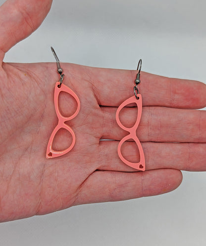 Image of a pair of fab sassy glasses earrings on a hand showing the size against a white background, they are two and a half finger widths length and one wide. They are sherbet pink and have black fixings.