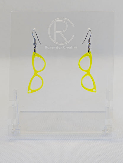 Shows a pair of acrylic earrings that have black ear wire hook fixings. They are fab sassy glasses with a heart earrings. They are neon yellow in colour and are against a white background on see through stand with the Ravenstor Creative logo on it.