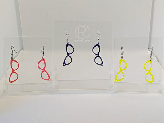 Shows three pairs of earrings that have ear wire hook fixings. They are fab sassy glasses earrings. The colours are pink, purple glitter and neon yellow. They are against a white background on see through stand with the ravenstor creative logo on.