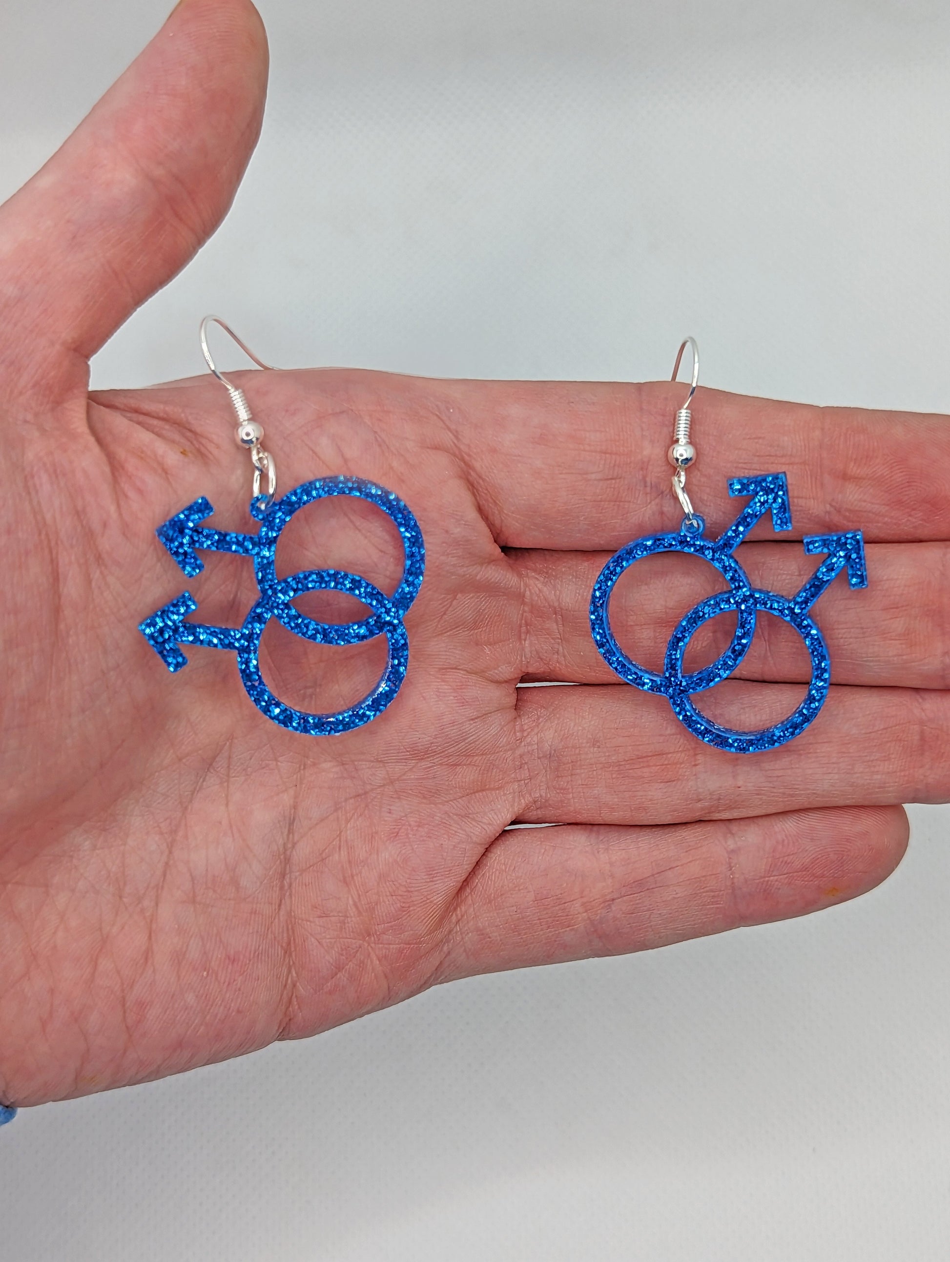 A pair of acrylic earrings in the shape of the Pride Double Male Gay LGBTQ symbol. They have ear wire hook fixings and are in Royal Blue Glitter. They are against a hand to show the size of 2 finger widths length and 2 wide.