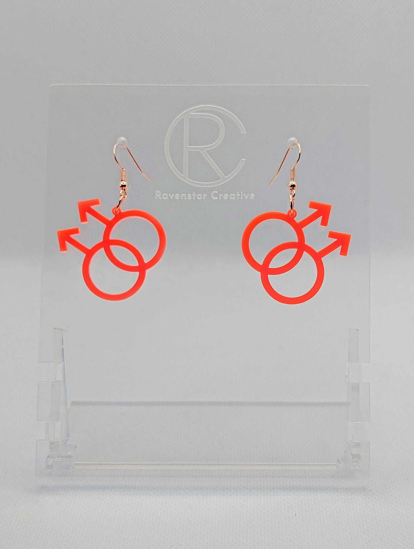 A pair of acrylic earrings in the shape of the Pride Double Male Gay LGBTQ symbol. They have Rose Gold ear wire hook fixings and the colour is Neon Pink.  They are on a clear stand with the Ravenstor Creative Logo on it against a white background.