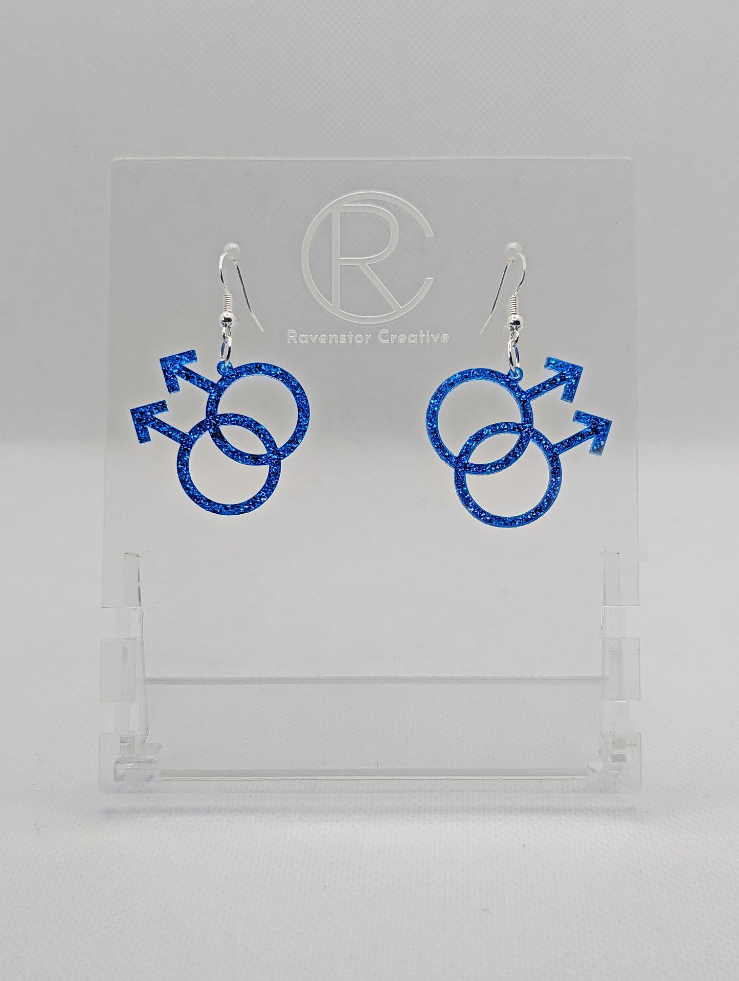 A pair of acrylic earrings in the shape of the Pride Double Male Gay LGBTQ symbol. They have Silver ear wire hook fixings and the colour is Ice Blue Glitter.  They are on a clear stand with the Ravenstor Creative Logo on it against a white background