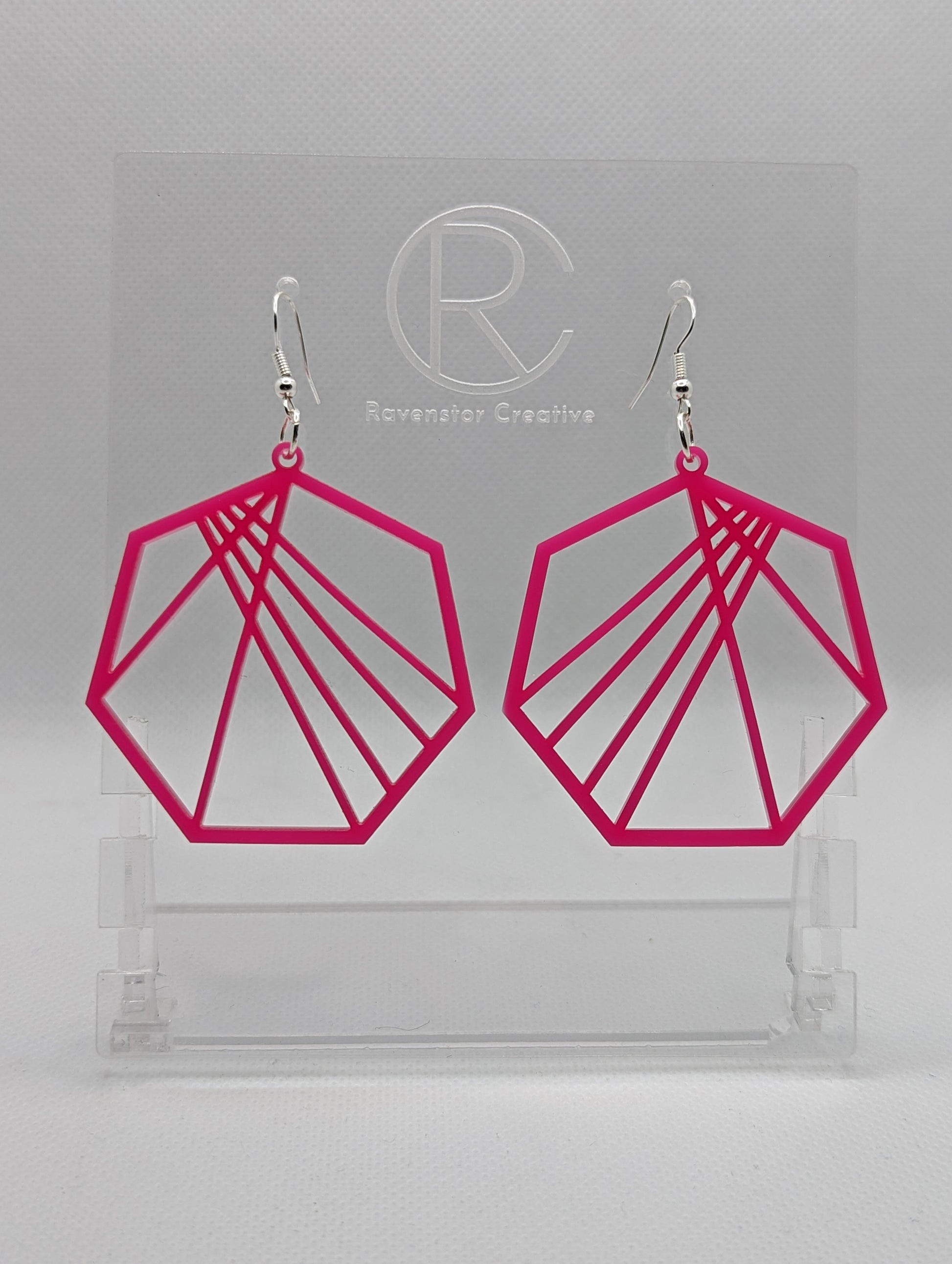 Shows a pair of earrings that have silver ear wire hook fixings. They are Geometric Heptagons with lines crisscrossing through the middle. They are made from Hot Pink Acrylic and are against a white background on see through stand.