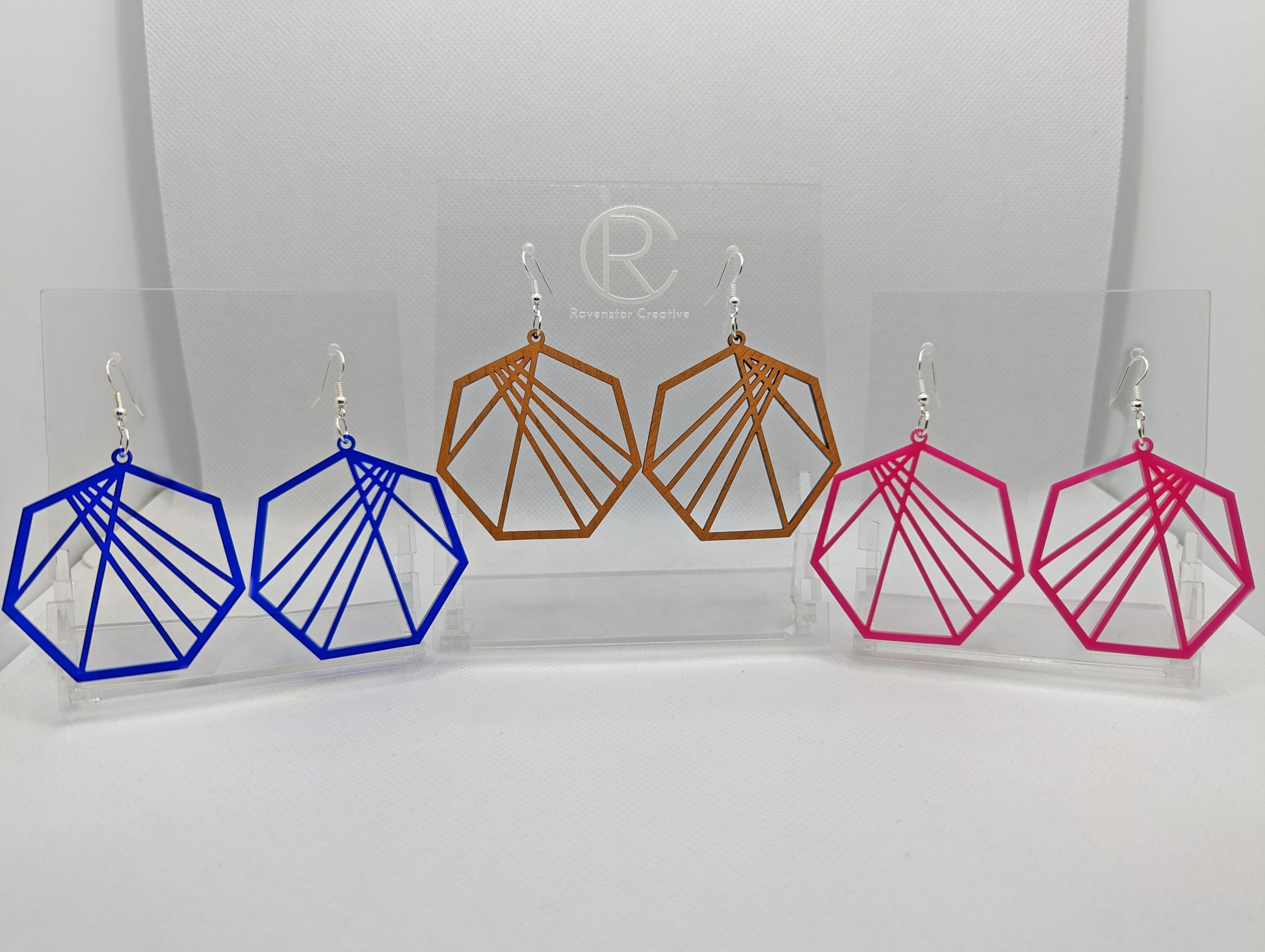 Shows three pairs of earrings that have ear wire fixings. They are Geometric Heptagon earrings with random crisscrossing lines. The colours are Translucent Blue, Cherry Wood and Hot Pink. They are against a white background on see through stands.