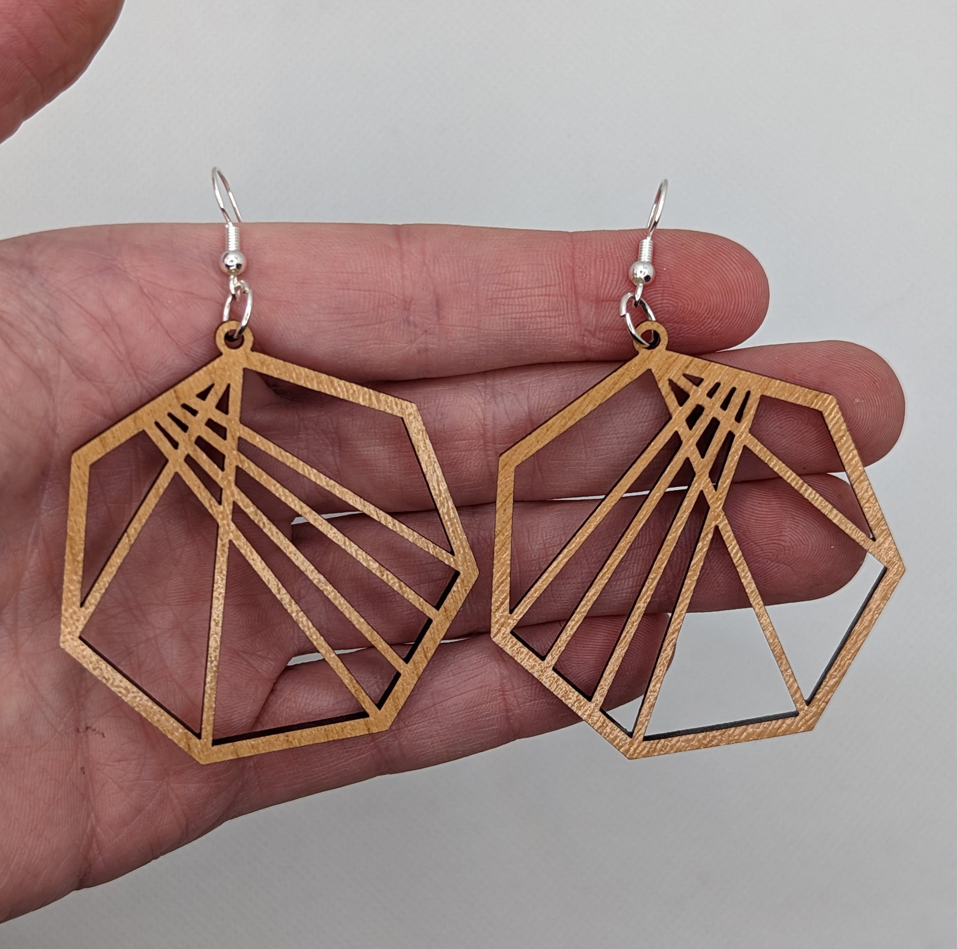 Image of a pair of Geometric Heptagon earrings on a hand showing the size against a white background. They are four finger widths in length and same again wide. They are made from Cherry Wood and have silver fixings.